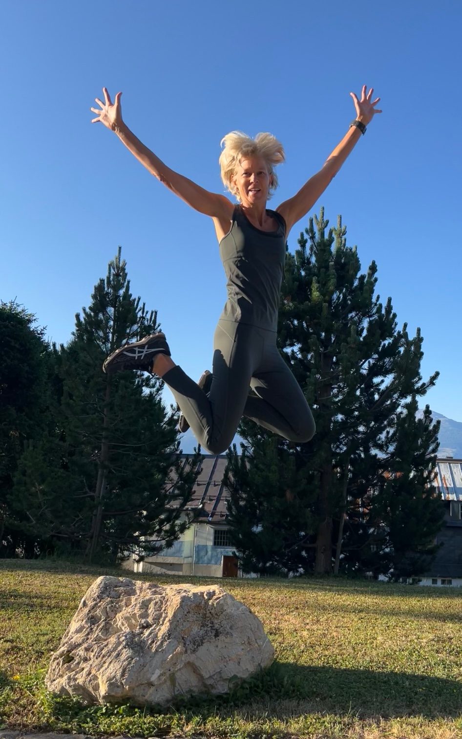 Laetitia Marchand saute, bras levés, au-dessus d’un rocher, devant des sapins et un ciel bleu