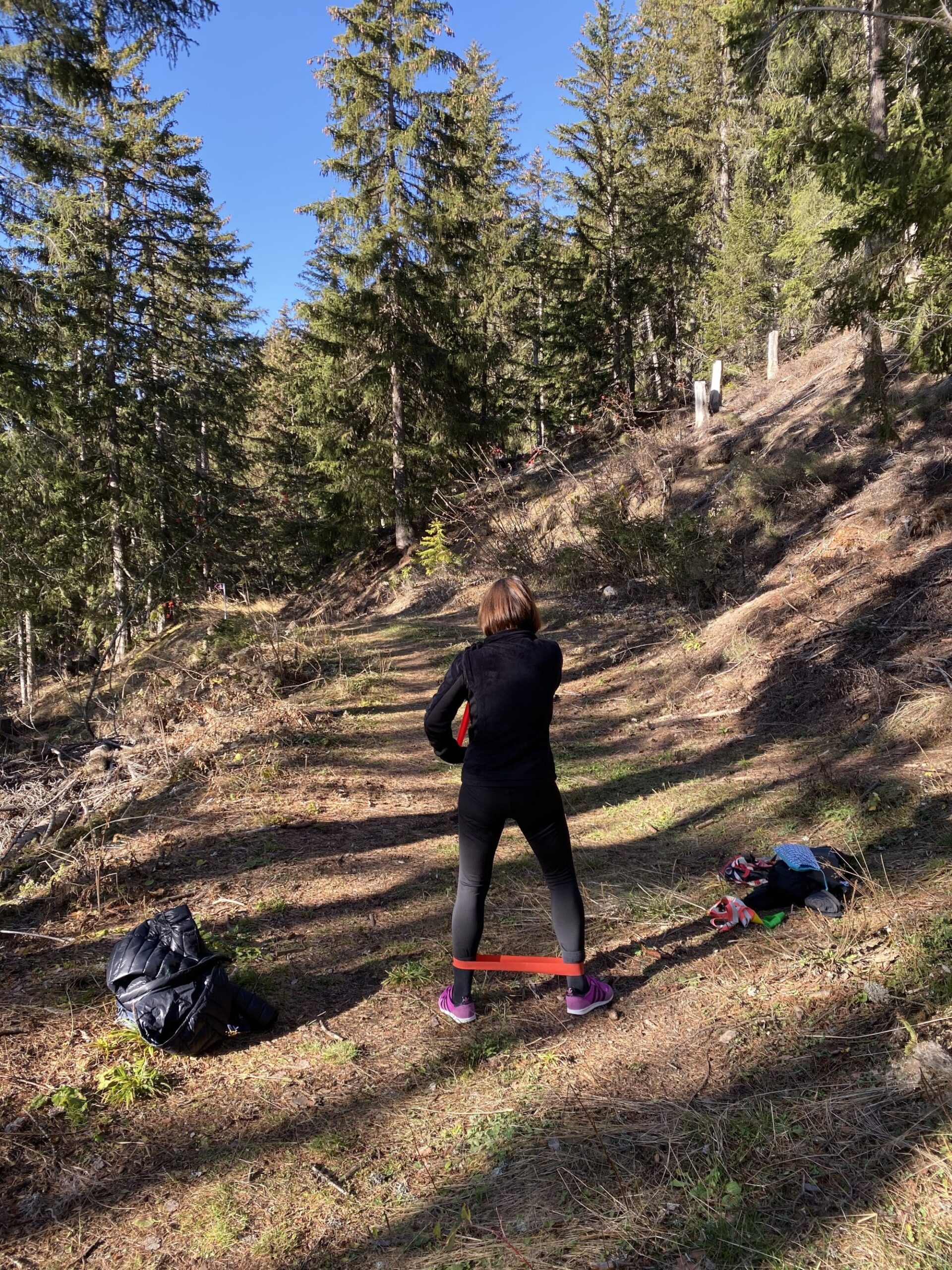Une personne de dos fait un exercice avec un élastique autour des chevilles, dans une clairière en forêt