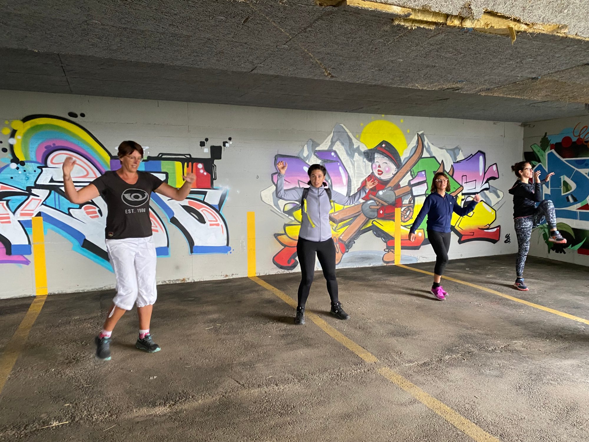 Quatre personnes font des exercices debout, à distance les unes des autres, devant un mur couvert de graffitis colorés
