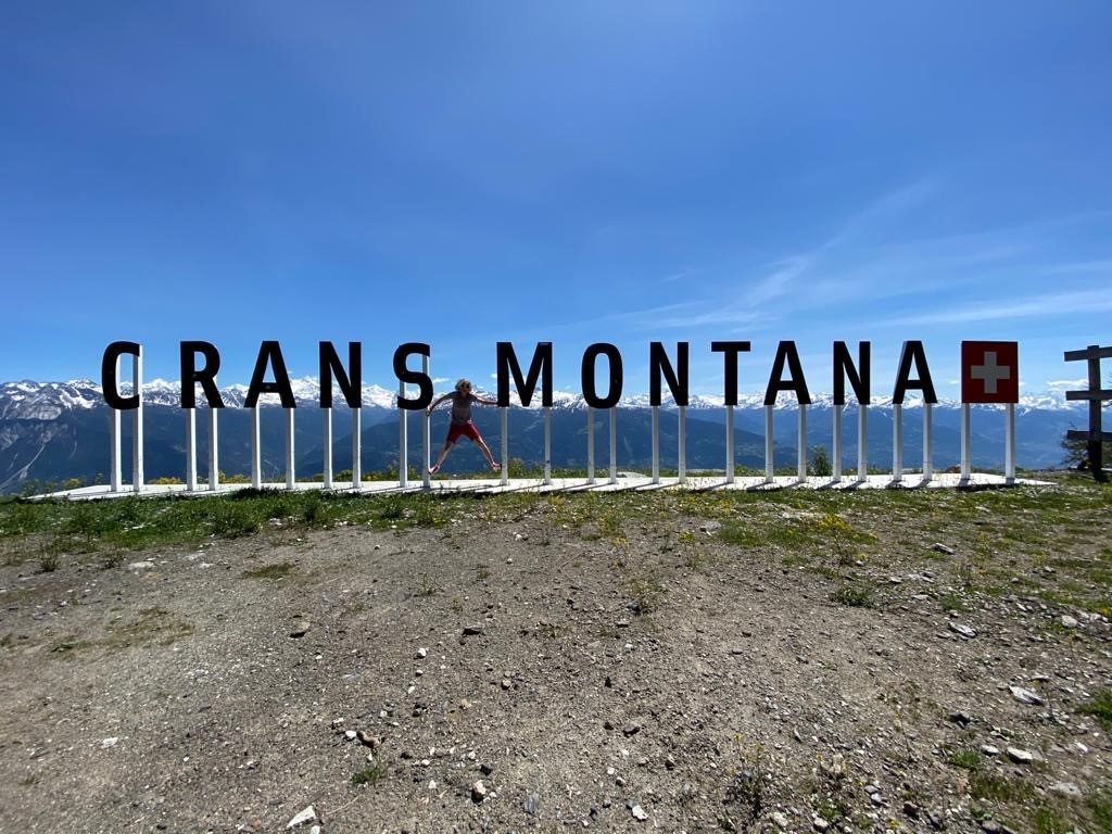 Les lettres géantes « Crans-Montana » et la croix suisse devant les Alpes ; une personne saute au milieu