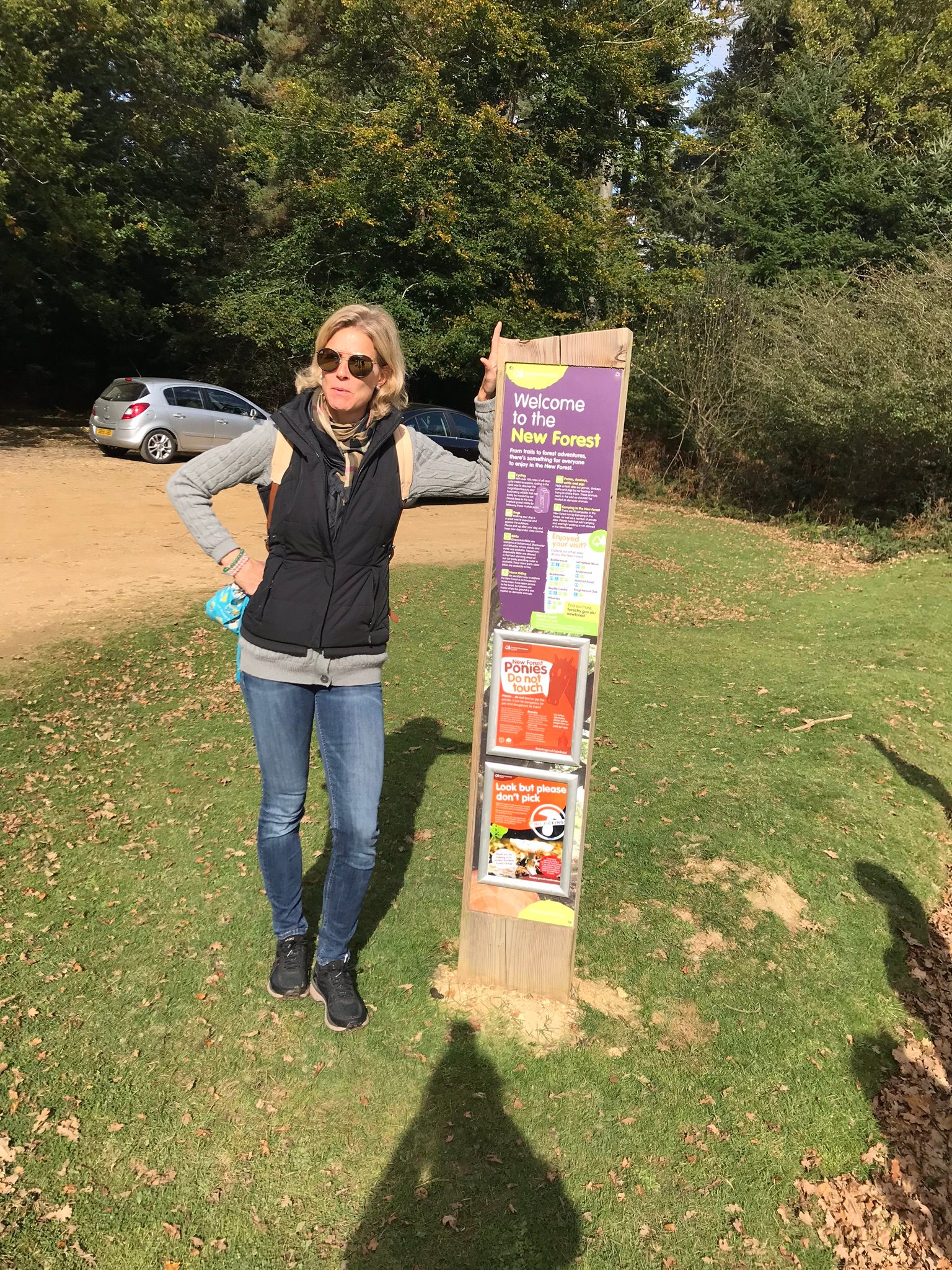 Une femme en tenue de sport pose à côté d’un panneau « Welcome to the New Forest », en lisière de bois