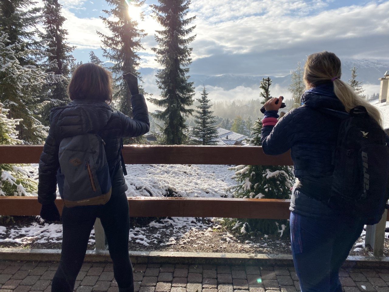 Deux personnes de dos s’étirent, un bras levé, devant une barrière en bois, face à des sapins saupoudrés de neige